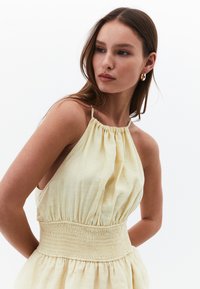 Žluté šaty s halter výstřihem, zdobené na krku a s jemně skládáním v pase. Volná sukně s měkkou texturou; minimalistický design bez vzorů.