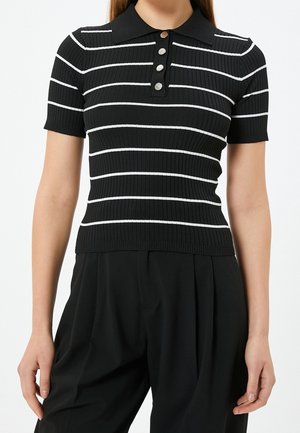Frau trägt ein schwarzes, kurzärmeliges, geripptes Poloshirt mit weißen horizontalen Streifen und silbernen Knöpfen, kombiniert mit schwarzen plissierten Hosen.