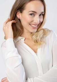 Witte, lichtgewicht blouse met lange mouwen en een knoopsluiting bij de hals. Model heeft golvend, schouderlang haar, glimlacht zachtjes en draagt oorbellen.