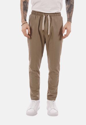 Hombre con pantalones casuales beige con cordón y zapatillas blancas, de pie con los brazos relajados a los lados.