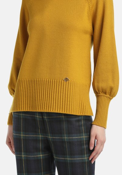 Mustard yellow sweater with a ribbed hem and sleeve cuffs, featuring a small silver logo detail. Worn with plaid dark blue and green pants.