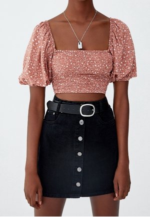Femme portant un crop top smocké rose à fleurs avec manches bouffantes, une jupe noire à boutons devant, une ceinture noire avec boucle argentée et un collier pendentif argenté.