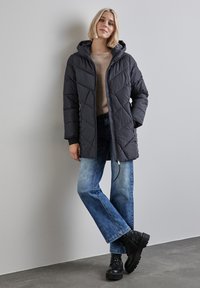 Černá péřová bunda s kapucí, s diamantovým vzorem, předním zipem a bočními kapsami, spojená se světle modrými rovnými džíny a robustními černými botami.