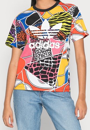 Kobieta w kolorowej koszulce Adidas z abstrakcyjnymi wzorami zwierząt w kolorach pomarańczowym, żółtym, różowym, niebieskim i czarnym, w połączeniu z jasnoniebieskimi jeansami.