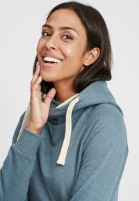 Sudadera con capucha azul de textura suave, con cuello de embudo y cordones de color crema. El modelo tiene el cabello oscuro y un fondo neutro.