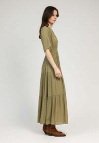 Robe maxi vert olive avec des manches courtes, à design superposé et taille cintrée. Portée avec des bottes de cow-boy marron, avec des coutures décoratives.