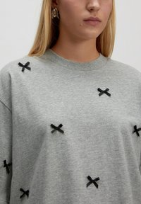 Grauer Sweatshirt mit großer, weicher Textur und strategisch platzierten, schwarzen Samtbows auf der Vorderseite für dekorative Details.