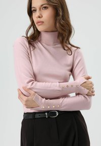 Lichtroze coltrui met lange mouwen en gouden knoopaccenten op de manchetten. Zacht materiaal met een aansluitend ontwerp. Zwarte riem zichtbaar.