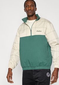 Πράσινο και μπεζ μπουφάν με φερμουάρ ως τη μέση, με σχέδιο πλέγματος, ελαστικές μανσέτες και χαμηλότερο τελείωμα με λάστιχο. Περιλαμβάνει μαύρη λογότυπη λεπτομέρεια στα παντελόνια.