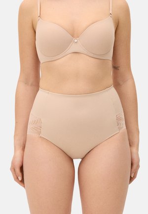 Mujer con sujetador color nude y bragas de talle alto con paneles laterales de encaje, de pie contra un fondo claro y liso.