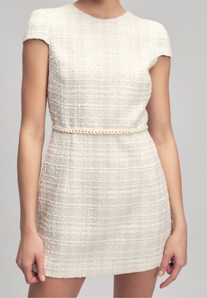 Kvinde iført en hvid tekstureret kortærmet kjole med et tyndt fletbånd om livet, foran en ensfarvet grå baggrund.