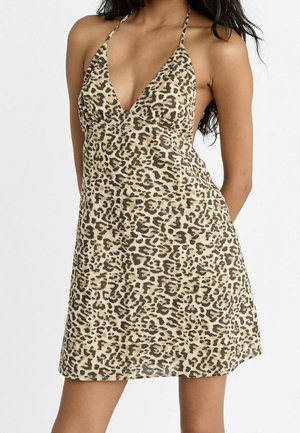 Femme portant une mini-robe sans manches, à col halter, imprimé léopard, sur un fond uni.