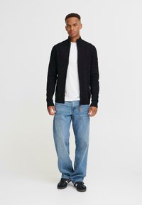 Chaqueta negra con cremallera sobre una camiseta blanca, combinada con jeans azul claros y zapatos negros. Diseño sencillo con puños ajustados y un look deportivo.