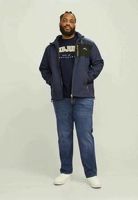 Homem de barba em pé com as mãos nos bolsos, vestindo um casaco azul-marinho, jeans azul-escuro, uma t-shirt gráfica azul-marinho e ténis brancos.
