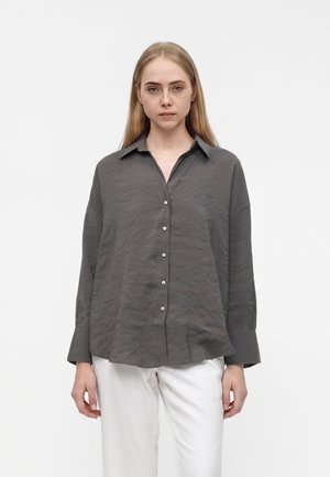 Jeune femme aux longs cheveux blonds portant une chemise grise ample boutonnée et un pantalon blanc, debout devant un fond clair uni.