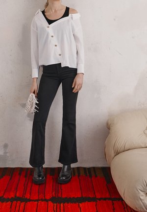 Femme portant une chemise blanche épaules dénudées boutonnée, un débardeur noir, un pantalon noir évasé, des bottes noires, tenant une pochette blanche tissée, debout sur un tapis rouge.