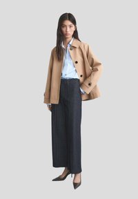 Beige kort kappa med stora knappar, blårandig skjorta och mörka vida jeans, kompletterat med spetsiga svarta klackar.