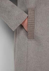 De hand van een persoon is gedeeltelijk gestoken in de geribbelde zak van een beige wollen jas met lange mouwen.