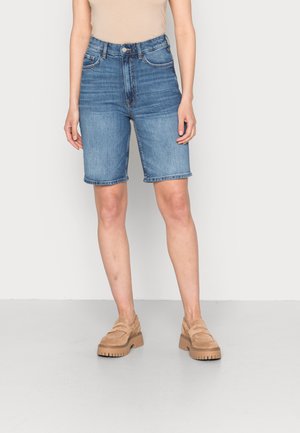 Person iført blå denimshorts i mellemlængde og lysebrune loafers, stående mod en ensartet lys baggrund.