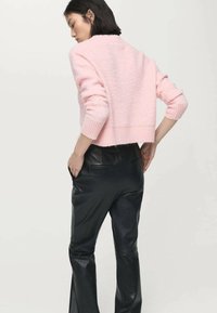 Vrouw die een lichtroze, pluizige trui en zwarte leren broek draagt, met haar handen in de zakken, met de rug naar de kijker en zijwaarts kijkend.