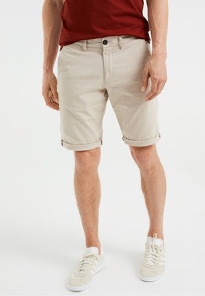 Mand iført beige opsmøgede shorts, hvide sneakers og en mørkerød skjorte står mod en ensfarvet baggrund.