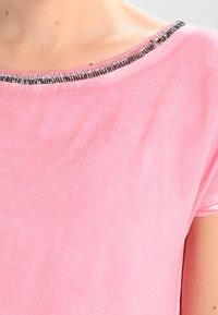 Nainen, jolla on päällä pehmeä vaaleanpunainen samettitoppi, jossa on pyöreä pääntie, joka on reunustettu hopea- ja mustilla helmill�ä.