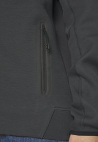 Grauer Kapuzenpulli mit Reißverschluss und glatter Textur, ausgestattet mit einer seitlichen Tasche und kontrastierenden Nähten entlang der Nähte.