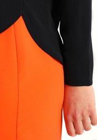 Person som bär en svart långärmad tröja med rundad nederkant över en ljusorange kjol eller byxor, handen avslappnad vid sin sida.