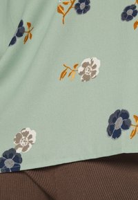 Květinová blůza v mátově zelené látce, s tmavě modrými, šedými a oranžovými květy a jemnými listy. Hladká textura, volný střih, zaoblený lem.