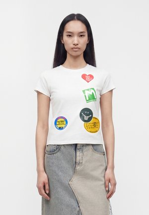 Femme portant un t-shirt blanc avec des patchs ronds et en forme de cœur colorés et une jupe en denim bicolore, debout devant un fond blanc.
