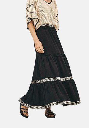 Falda larga negra con volantes y ribete trenzado blanco en el dobladillo, combinada con una blusa de punto beige suelta que presenta detalles en negro y un escote en V.