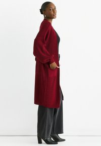 Long red cardigan with a ribbed texture, oversized sleeves, open front, and side pockets, paired with black wide-leg pants and heeled boots.