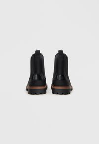 Par de botines de cuero negro con suelas medias marrones y suelas robustas negras, vistos desde atrás sobre un fondo claro.