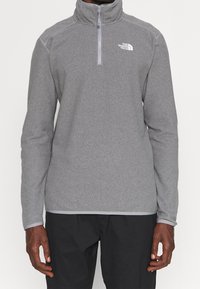 Grå zip-up fleece-tröja med hög krage, featuring en logga på bröstet och långa ärmar. Slät textur och atletisk passform.