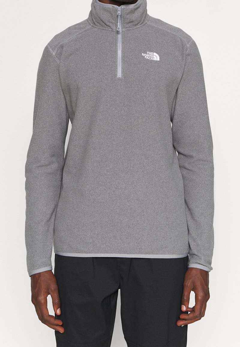 Grå zip-up fleece-tröja med hög krage, featuring en logga på bröstet och långa ärmar. Slät textur och atletisk passform.