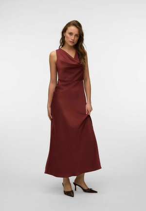 Femme portant une robe longue en satin couleur rouille sans manches et des chaussures à talons aiguilles marron pointues, debout devant un fond clair uni.