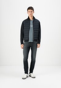 Padded black jacket with a zip front, grey shirt underneath, dark jeans, and white/grey sneakers. Smooth texture, minimal design.