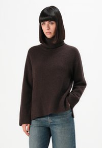 Femme aux cheveux sombres et lisses portant un pull col roulé marron foncé et un jean bleu, se tenant avec une main dans la poche, sur fond blanc.