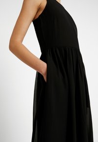 Robe noire sans manches avec taille froncée, jupe à superposition transparente et poche latérale, portée par une personne avec un bras plié à la taille.
