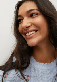 Jasnoniebieski sweter z dzianiny z warkoczowym wzorem i kwiatową haftem. Modelka ma długie ciemne włosy i widoczny uśmiech, na białym tle.