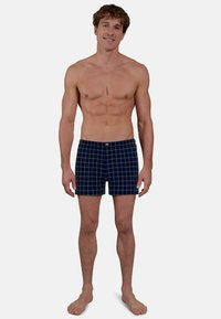 Lachende man zonder shirt die blootsvoets staat, met blauw en groen geruite boxershorts, tegen een effen lichte achtergrond.