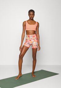 Sutiã desportivo rosa combinado com calções de padrão floral vibrante. O tecido parece elástico. A modelo está em pé sobre um tapete de yoga verde.