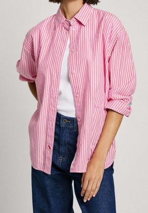 Persoon draagt een roze en rode verticale gestreepte overhemd over een witte top met opgerolde mouwen en een blauwe spijkerbroek.