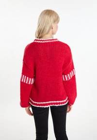Czerwony sweter z dzianiny z białymi akcentami. Luźny krój, ryżowy splot oraz kontrastowe paski na rękawach i u dołu. Widok z tyłu.