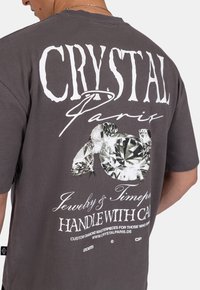 Šedé bavlněné tričko s velkým bílým grafickým potiskem diamantů a textem "CRYSTAL Paris Jewelry & Timepieces" na zádech.