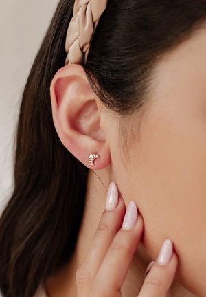 Ohrringe aus roségold in Form eines Halbmonds, mit einer strukturierten Oberfläche und einem kleinen Schmucksteinakzent. Haare mit einem leichten Haarband gestylt.