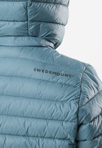 Ljusblå vadderad jacka med horisontellt quiltat mönster, fastsydd huva och "SWEDEMOUNT"-logotyp på vänster axel. Mjuk textur.