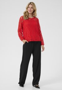 Red lace blouse with long sleeves, scalloped neckline, layered texture. Paired with black wide-leg trousers and black flat shoes.