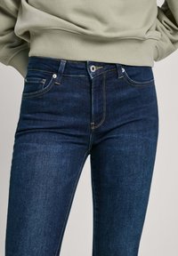 Jeans de mezclilla azul oscuro con un diseño ajustado. Las características incluyen un estilo clásico de cinco bolsillos, costuras doradas y un cierre frontal con botón.