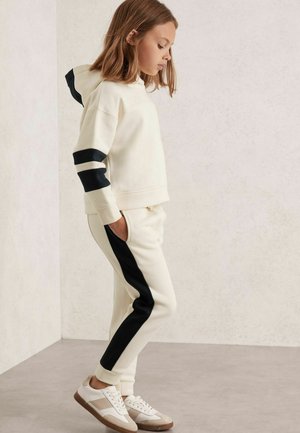 Creme hoodie med sorte accentstriber, parret med creme joggers med sorte sidepaneler. Hvite sneakers med beige accenter.